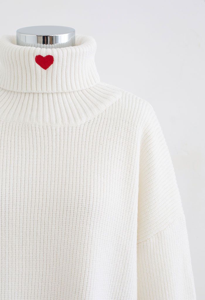 Front view of a cozy white turtleneck crop sweater with an embroidered red heart near the collar.