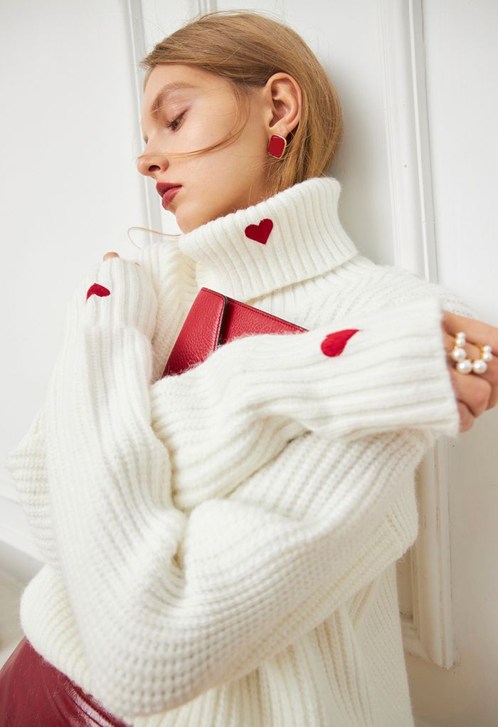 Side view of a model in a white turtleneck sweater with red heart details, paired with a red clutch.