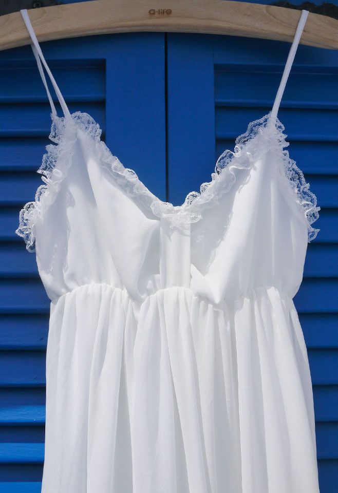 Close-up of the lace-trimmed neckline and straps of a white chiffon maxi dress, hanging on a wooden hanger.