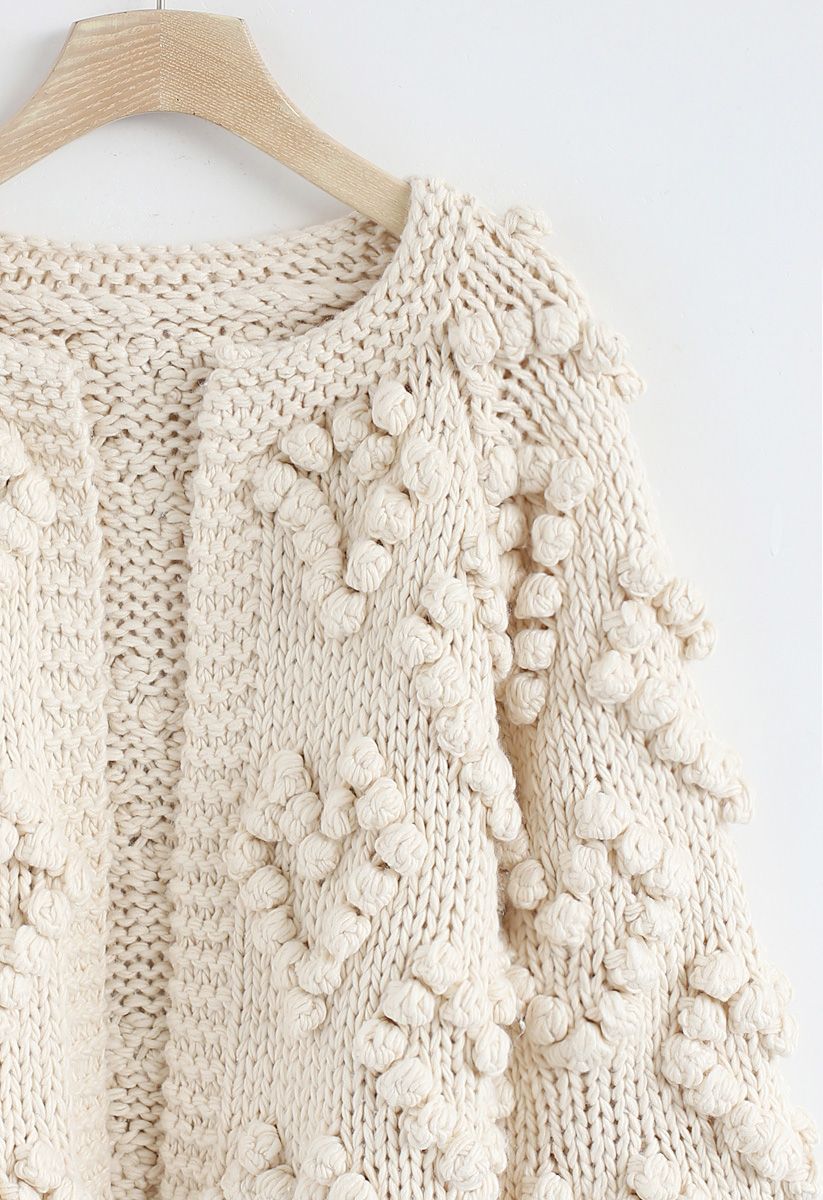 Detail shot of an ivory cardigan with intricate knit patterns and textured bobbles, hanging on a wooden hanger.