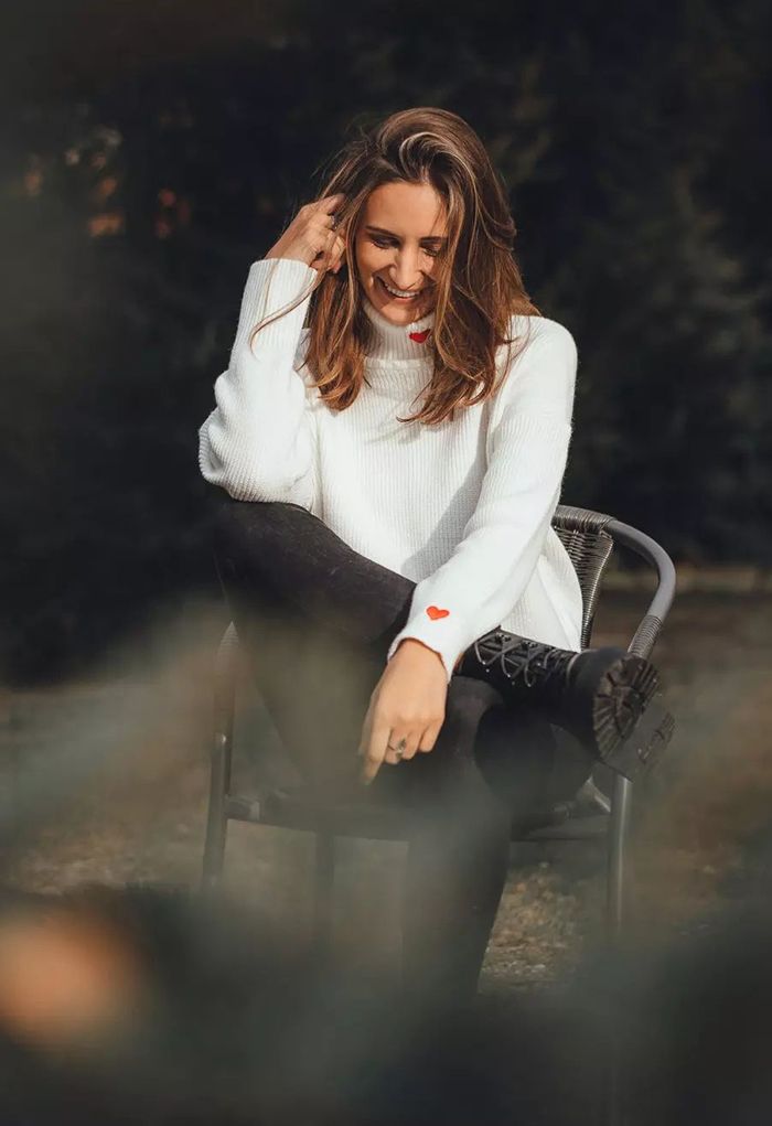 A woman sitting outdoors in a white turtleneck sweater with red heart embroidery, exuding a relaxed vibe.