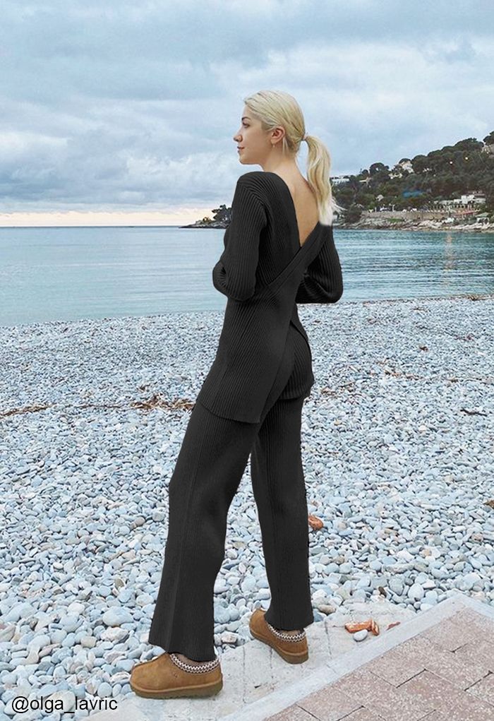 A person standing on a pebble beach wearing a black knit outfit, looking out at the ocean.